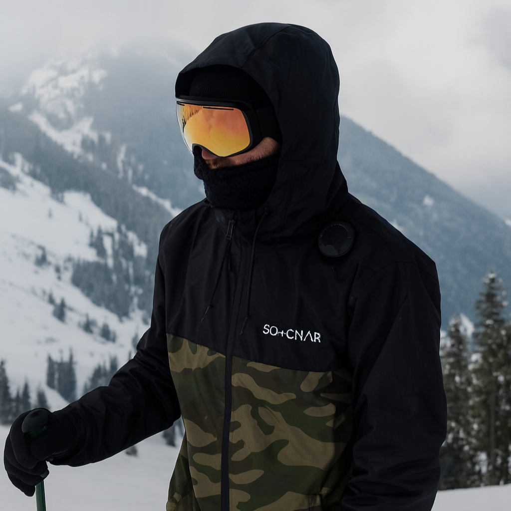 Person wearing a black and camouflage jacket with goggles, standing on a snowy mountain.